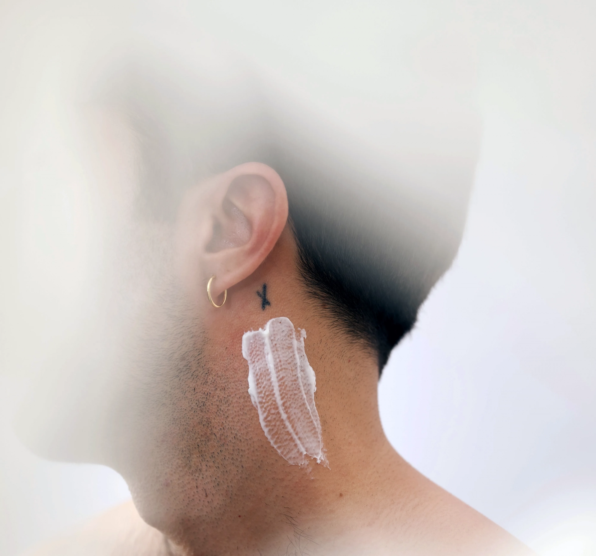 El cuello de un hombre de perfil con un tatuaje de una X y una muestra de la crema facial Mousse Nutritivo de Piel·e Cosmetics; muestra de cosmética en un hombre